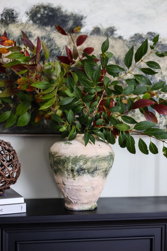 rustic looking vase filled with green branches on a mantle.